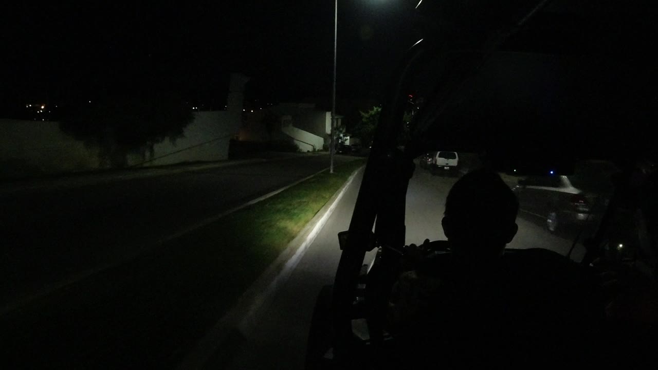 The jeep-mounted view continues along a dark Los Cabos street, showing a pale wall, roadside grass, a parked car, and foreground silhouettes without identifiable faces.