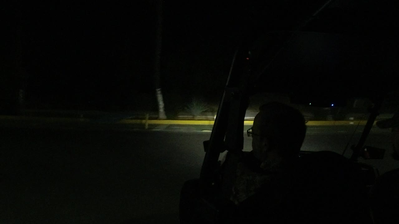 The road continues ahead in Los Cabos at night, with the vehicle frame and passenger silhouette at the right and small curbside lights visible in the distance.