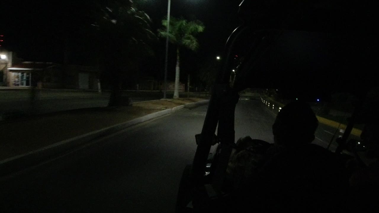 The Wrangler continues along a dark Los Cabos street, with a passenger silhouette centered low in frame, roll bars at both sides, and a lit building and palm tree near the road.