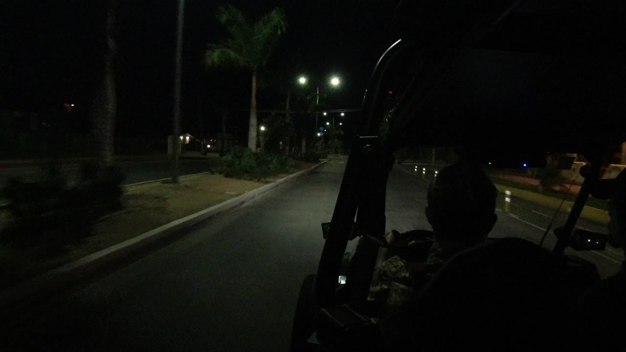 The Los Cabos night street remains ahead of the open Wrangler, with the driver in shadow at right, double streetlights, curbside plants, and low roadside structures in the distance.