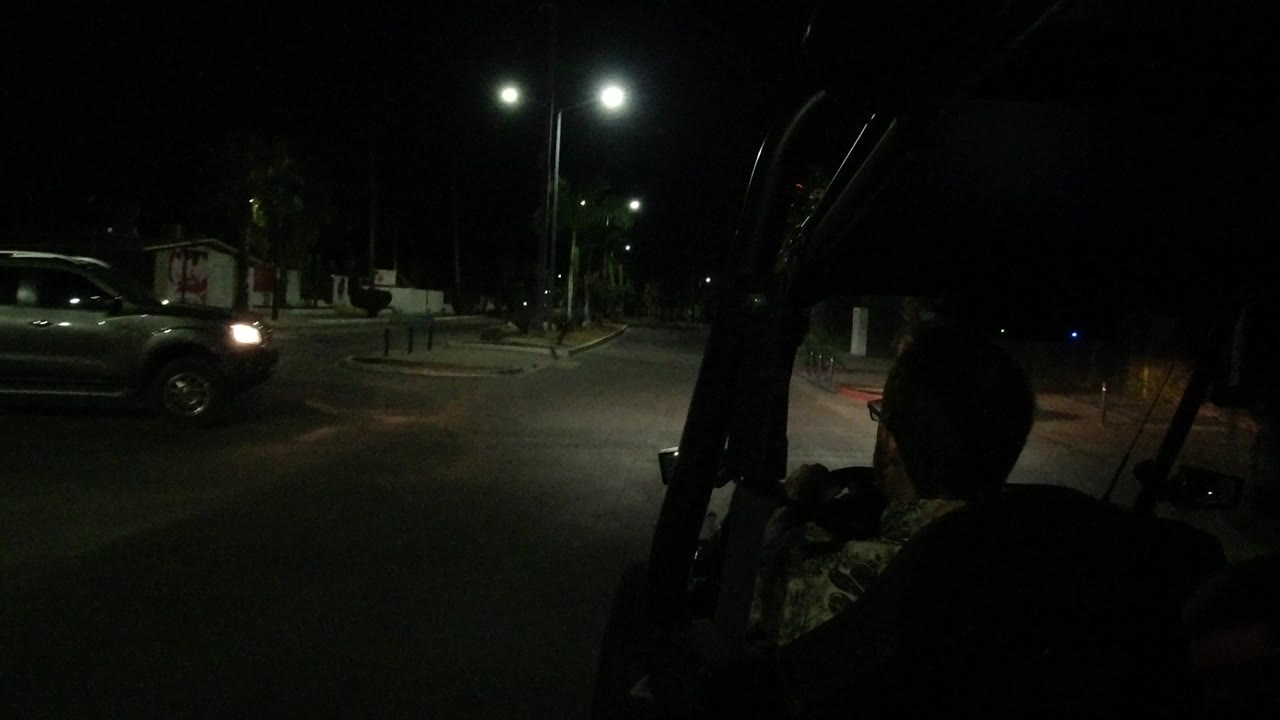 Night street seen from inside an open vehicle with a person silhouetted in front.