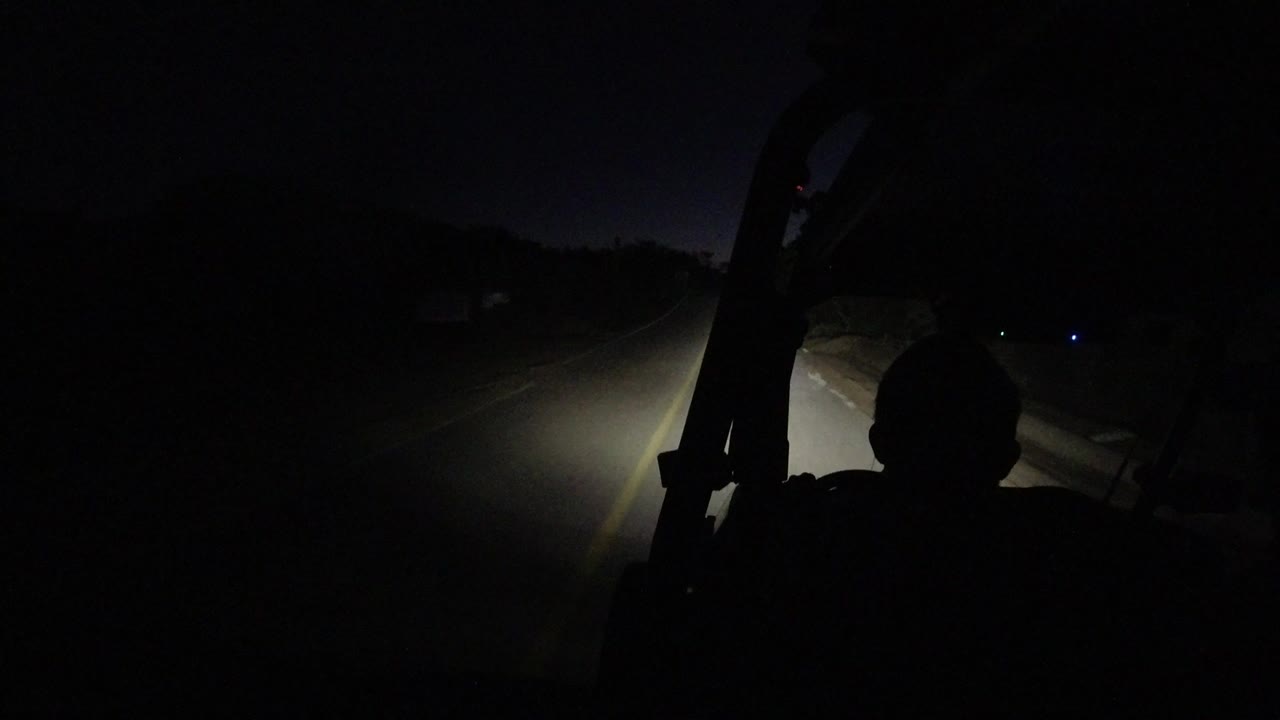 Opening frame of a Los Cabos night drive, with headlights lighting the road edge and a dark vehicle interior silhouette in the foreground, no identifiable people visible.