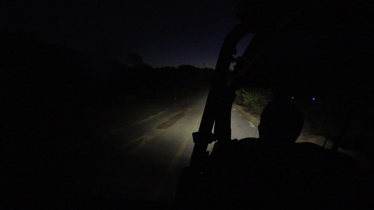 Side-mounted view in Los Cabos, Mexico with the jeep frame silhouetted at right and headlights lighting the left edge of a dark unlit road, with no visible people.