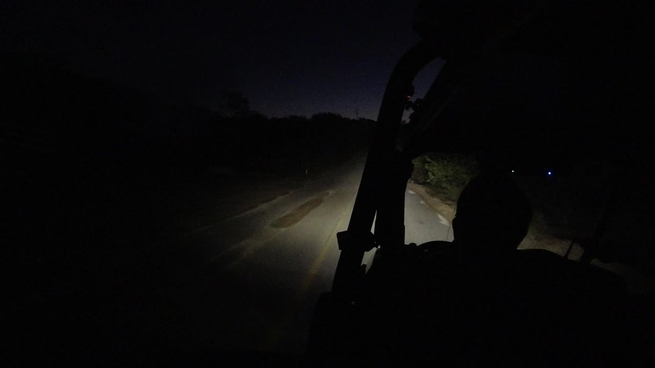 A dark Los Cabos road sits ahead of the side-mounted jeep view, with the headlight beam spreading over pavement and roadside vegetation, with no visible people.