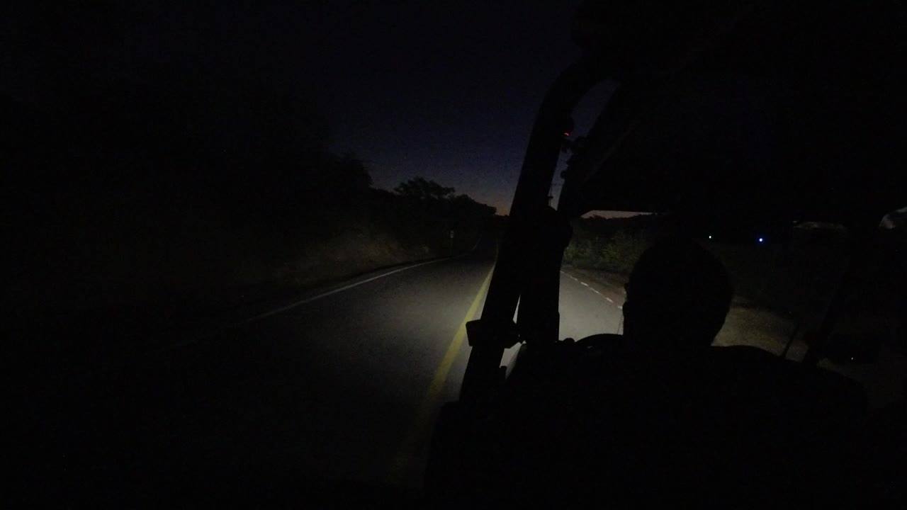 Night road view in Los Cabos, Mexico, seen from inside a vehicle as headlights light the lane and a dark seated silhouette appears in the foreground.