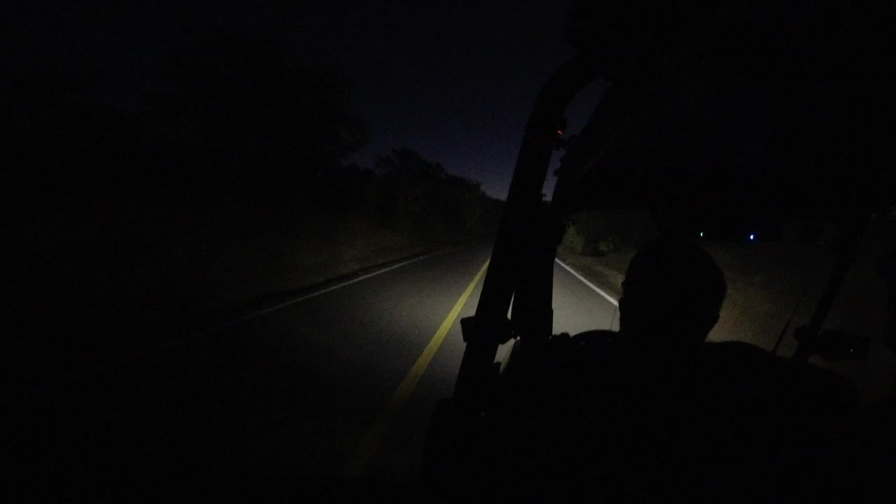 One second in, the Los Cabos road curves through darkness as the vehicle headlights catch the lane edge and the foreground person remains silhouetted.
