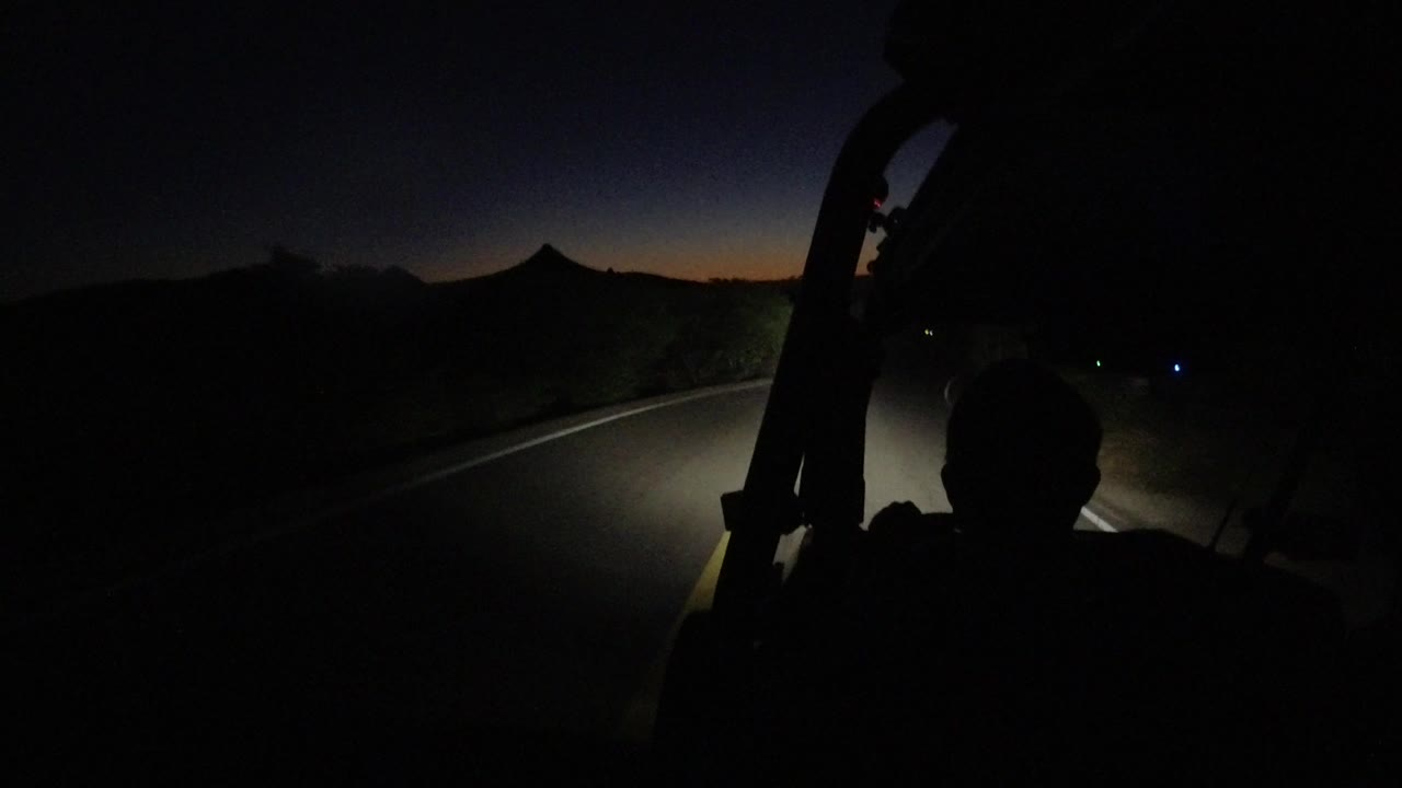 A forward-facing Los Cabos road view shows the headlight pool on pavement, a shadowed roadside, a faint orange horizon, and a seated silhouette without visible facial features.