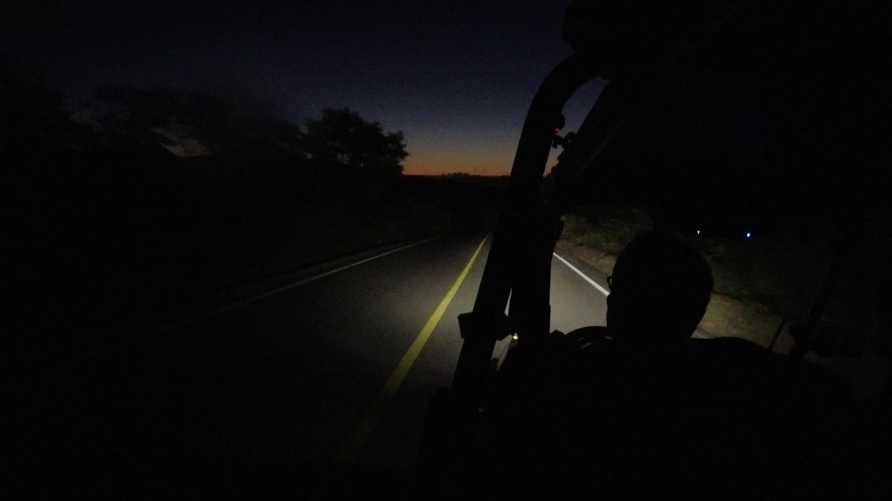 Los Cabos road at 0:01 with the vehicle slightly farther along the lane, headlights lighting the pavement, a faint blue roadside light, and a person silhouetted at lower right.