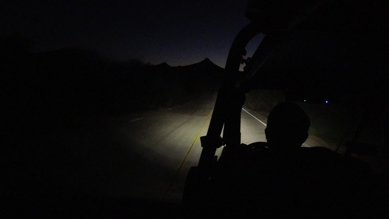 A silhouetted buggy driver in Los Cabos, Mexico, faces a narrow dark road where the headlights reveal the lane edge and roadside vegetation.