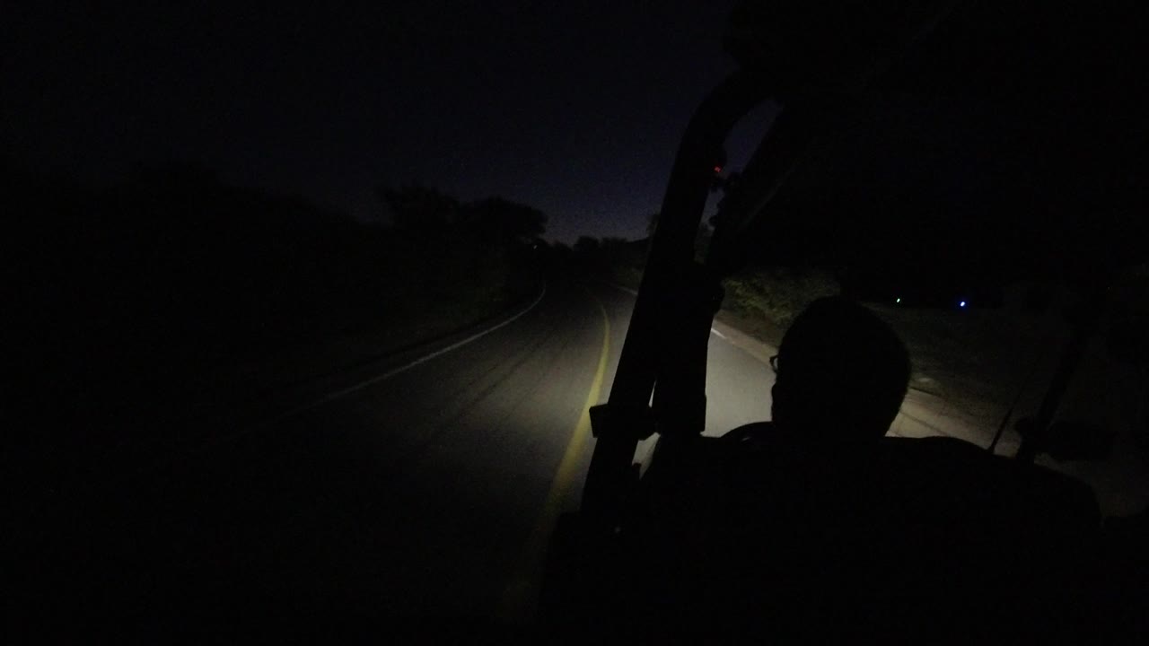 At 0:01, the Los Cabos road curves through darkness with the vehicle structure and a seated silhouetted person blocking part of the right foreground.