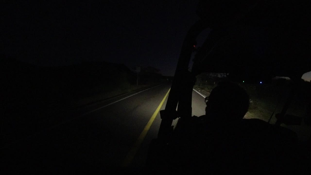 The forward view from the vehicle shows a yellow center line, roadside vegetation, and scattered distant lights in Los Cabos, Mexico, with the occupant remaining a dark silhouette.