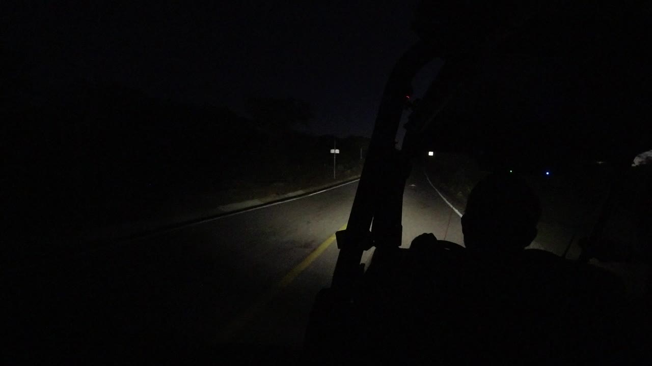At 0:00 in Los Cabos, Mexico, headlights illuminate the centerline and shoulder of a dark highway while a foreground silhouette sits low in frame.