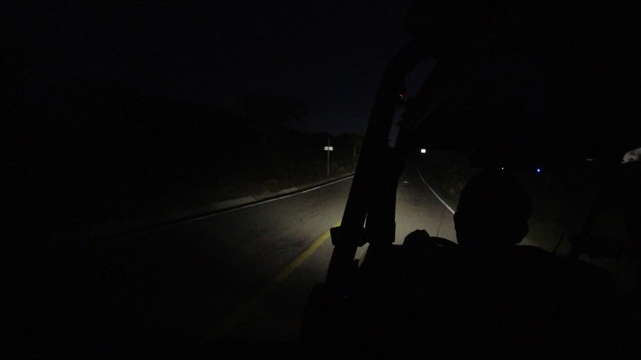 At 0:01 in Los Cabos, Mexico, the road extends into darkness with a silhouetted rider or passenger shape at the lower center and a small blue light ahead.