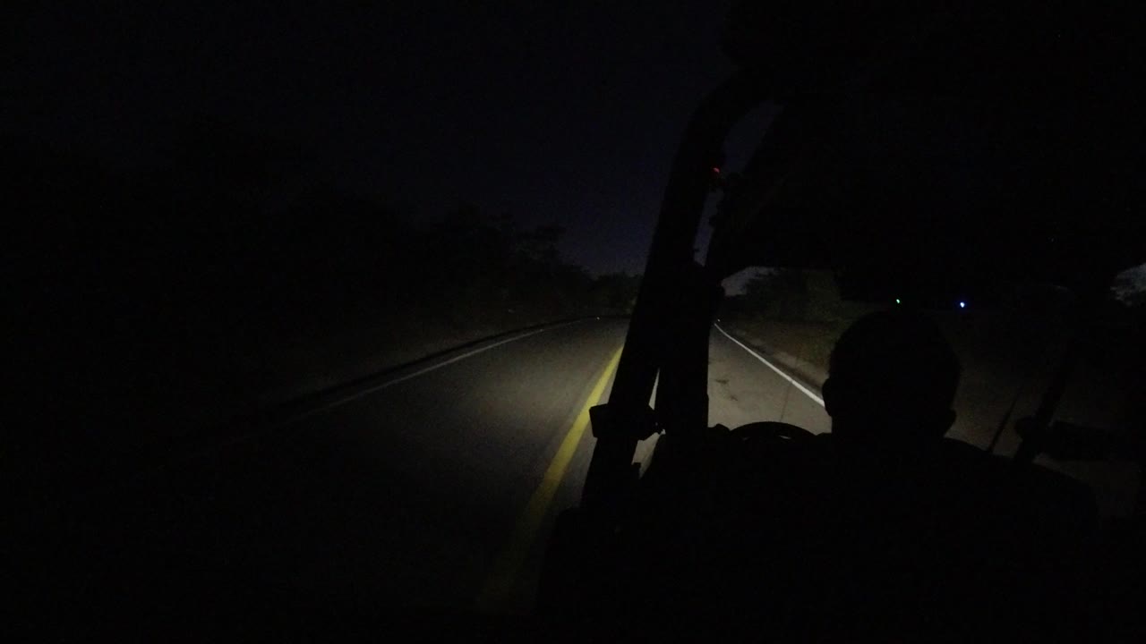 A dark Los Cabos road seen from inside a vehicle at night, with headlights illuminating the yellow center line and a silhouetted occupant in the foreground.