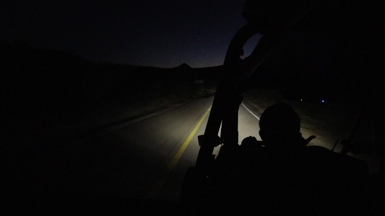 Dark road ahead lit by vehicle headlights with a silhouetted passenger in front.