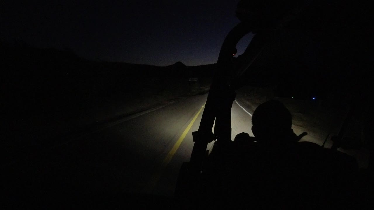 The vehicle continues along a dark Los Cabos road, with the yellow center line bright in the headlights and a silhouetted person seated in the lower frame.