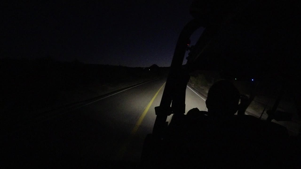 At 0:00 in Los Cabos, Mexico, a dark vehicle interior frames a night road, with a driver silhouette on the right and the headlamp beam lighting the yellow line and roadside grass.