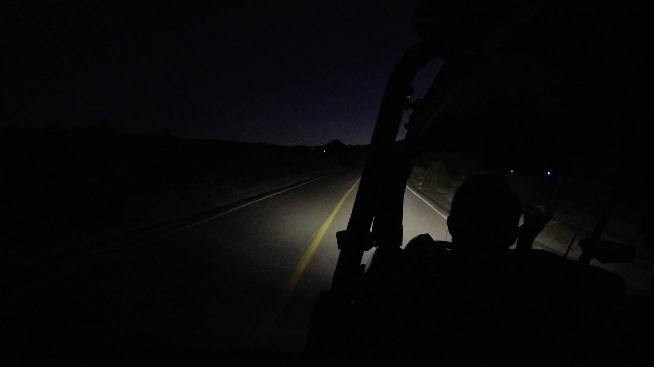 At 0:01 in Los Cabos, Mexico, the headlamp beam falls farther along the road while the driver's head remains a dark foreground shape beside the vehicle frame.