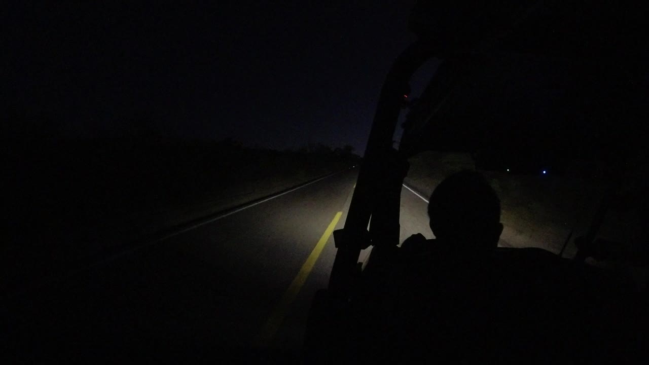 A Los Cabos nighttime road is lit by jeep headlights, with a dark human silhouette in the foreground and brush barely visible along the roadside.
