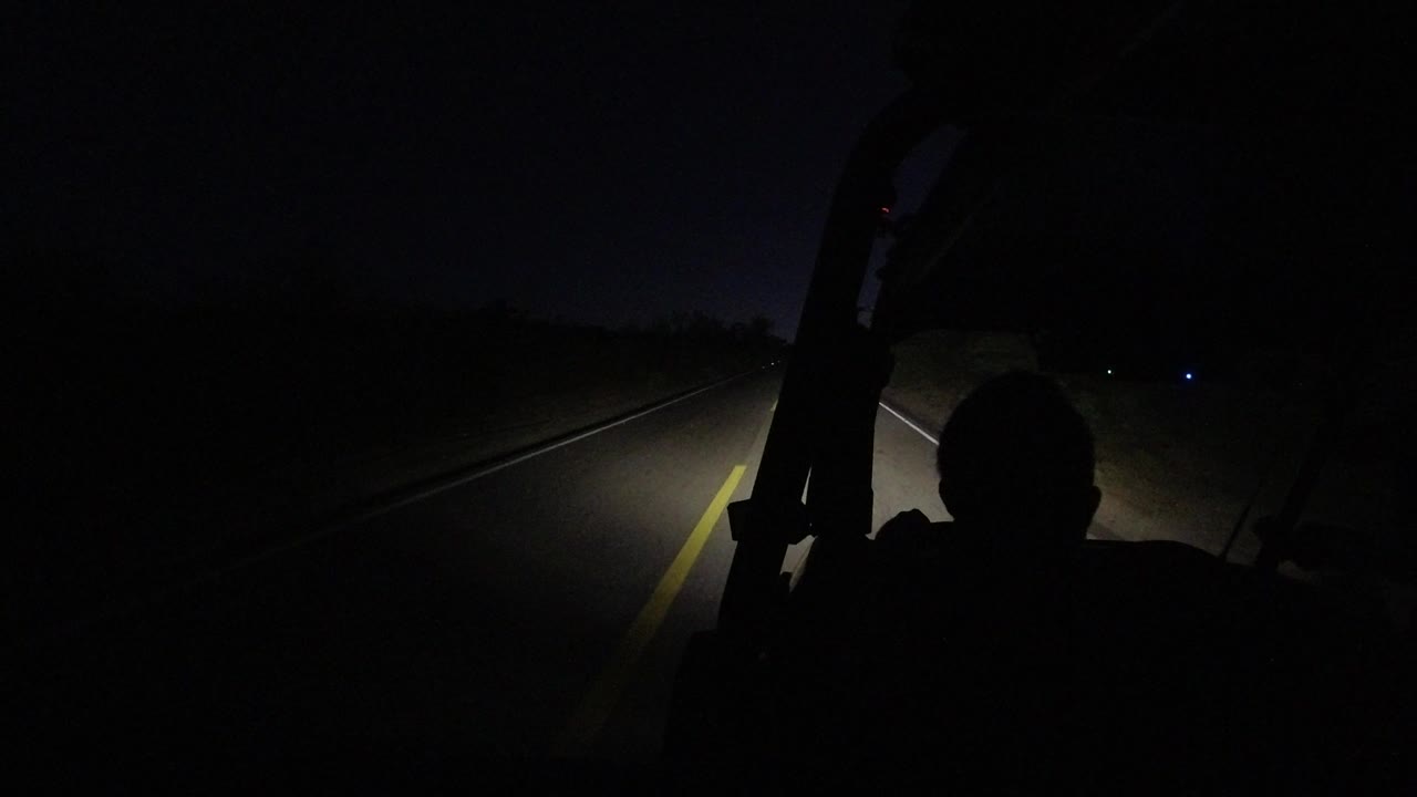 The jeep headlights sweep across a dark Los Cabos road, with the silhouetted back of a person low in frame and a faint roadside horizon ahead.