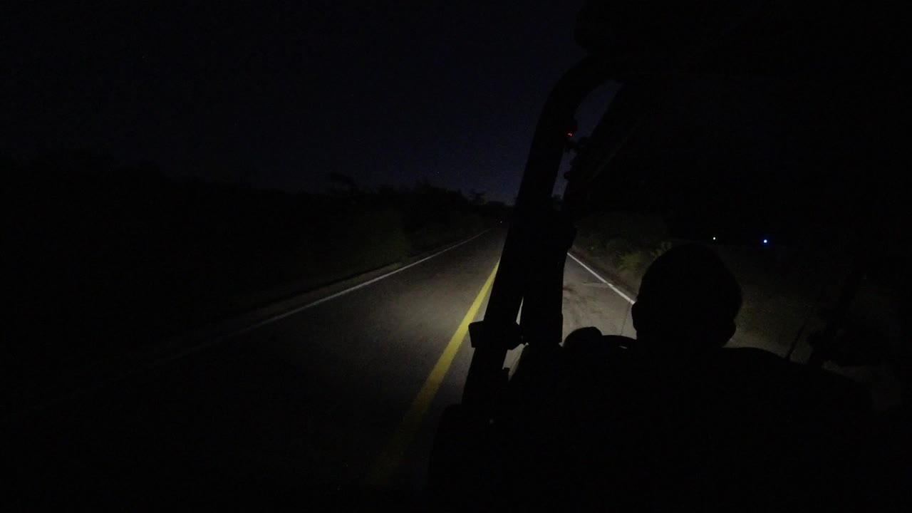 At 0:01, the headlight beam reaches farther down the Los Cabos road while the vehicle interior and a foreground head remain nearly black.