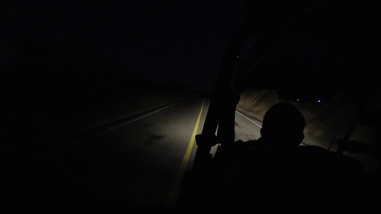 At 0:01, the Los Cabos road remains mostly black, with the yellow lane line brighter on the right and the driver silhouette centered low in frame.