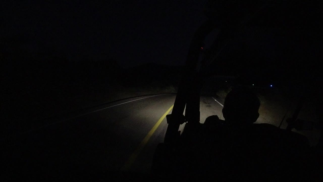 Opening frame of a Los Cabos night drive with a yellow road line, headlight beam, windshield frame, and a dark seated silhouette in the foreground.