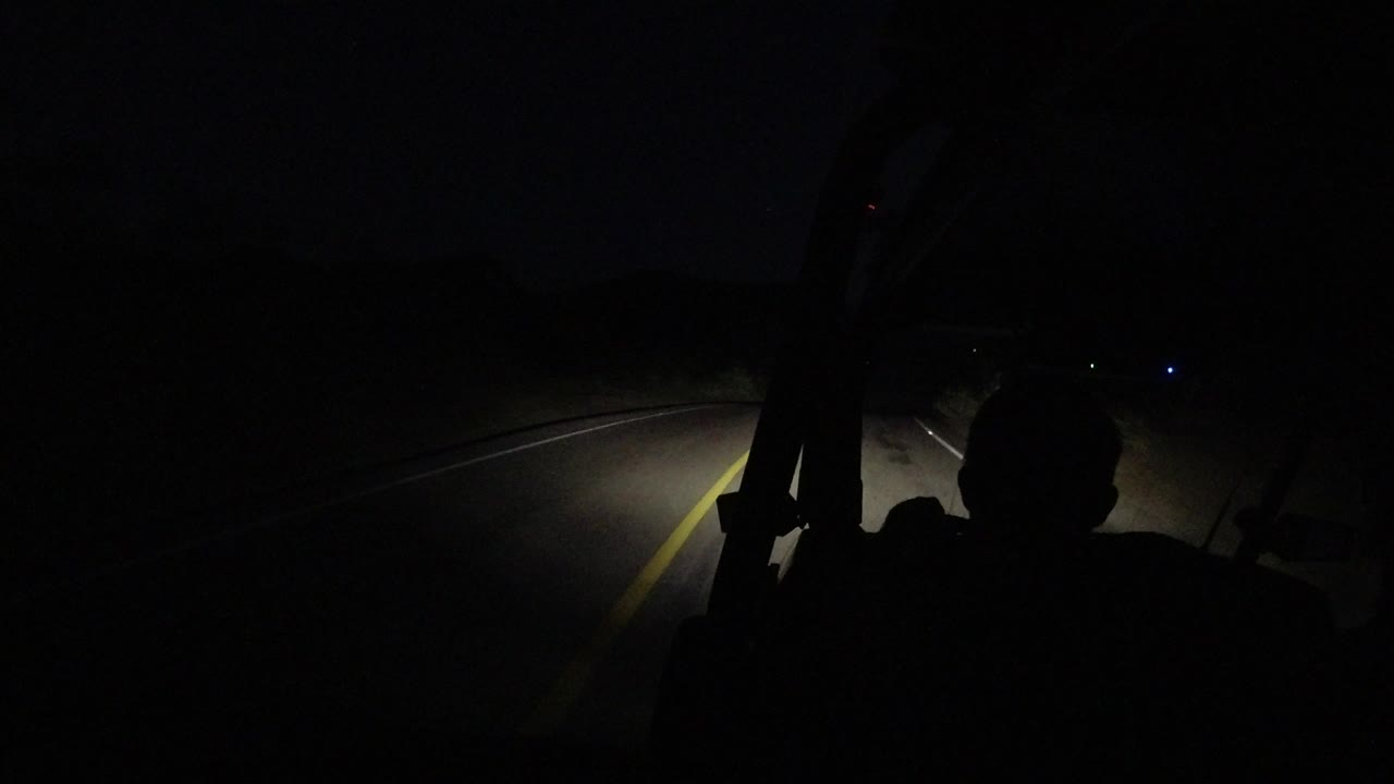 A Los Cabos road curves ahead in darkness, with the jeep frame and a seated foreground silhouette blocking part of the headlight-lit lane.