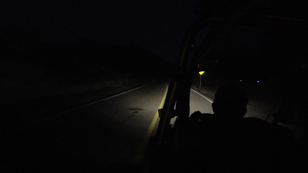 In Los Cabos, Mexico, the road remains mostly black except for the headlight cone, with the bus interior framing the view and a silhouetted person centered low in frame.