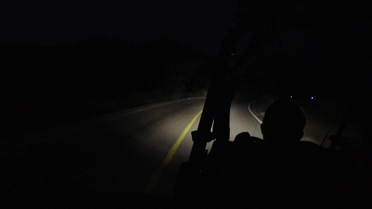 At 0:00, a dark Los Cabos road curves ahead under vehicle headlights, with a yellow lane line and cabin silhouettes in the foreground, no visible people.