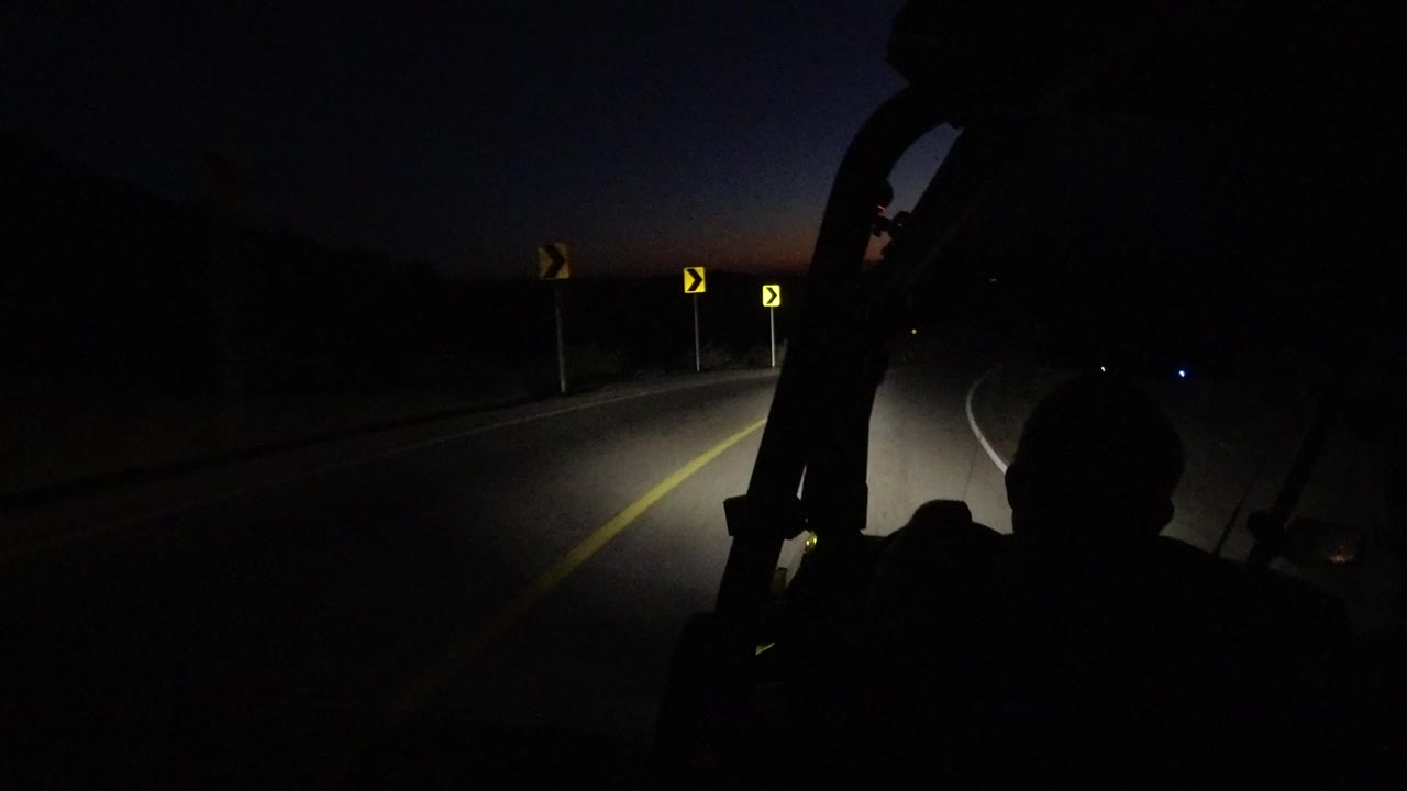 A dark Los Cabos road curves ahead under headlights, with yellow curve signs, a thin orange horizon, foreground vehicle silhouettes, and no visible people.