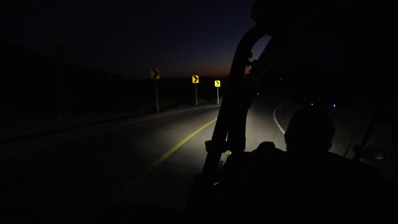 The road bends through darkness in Los Cabos, with two yellow curve markers ahead, headlight spill across the lane, and no visible people.