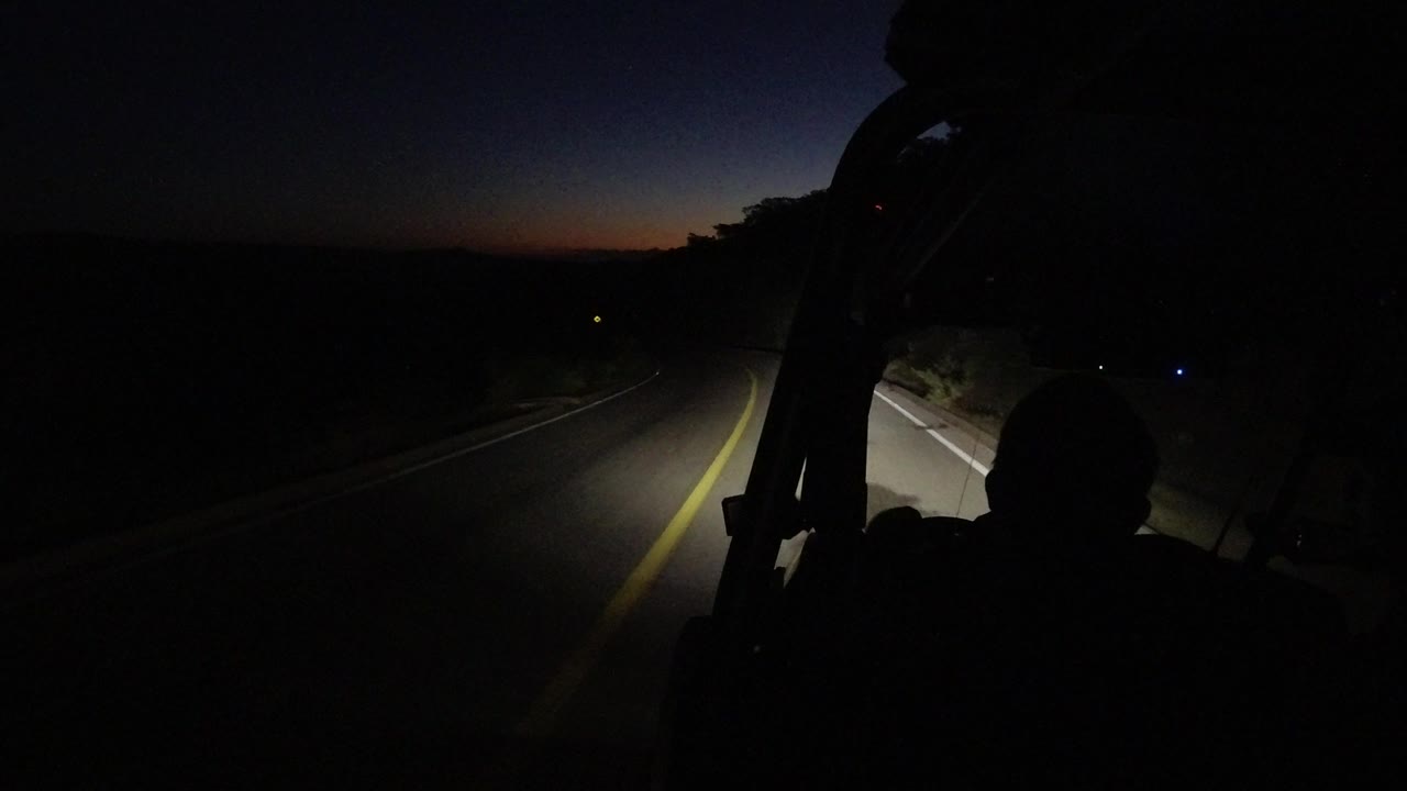 At 0:01 in Los Cabos, Mexico, the headlight beam tracks along the yellow road line while the foreground remains mostly black around a seated silhouette.