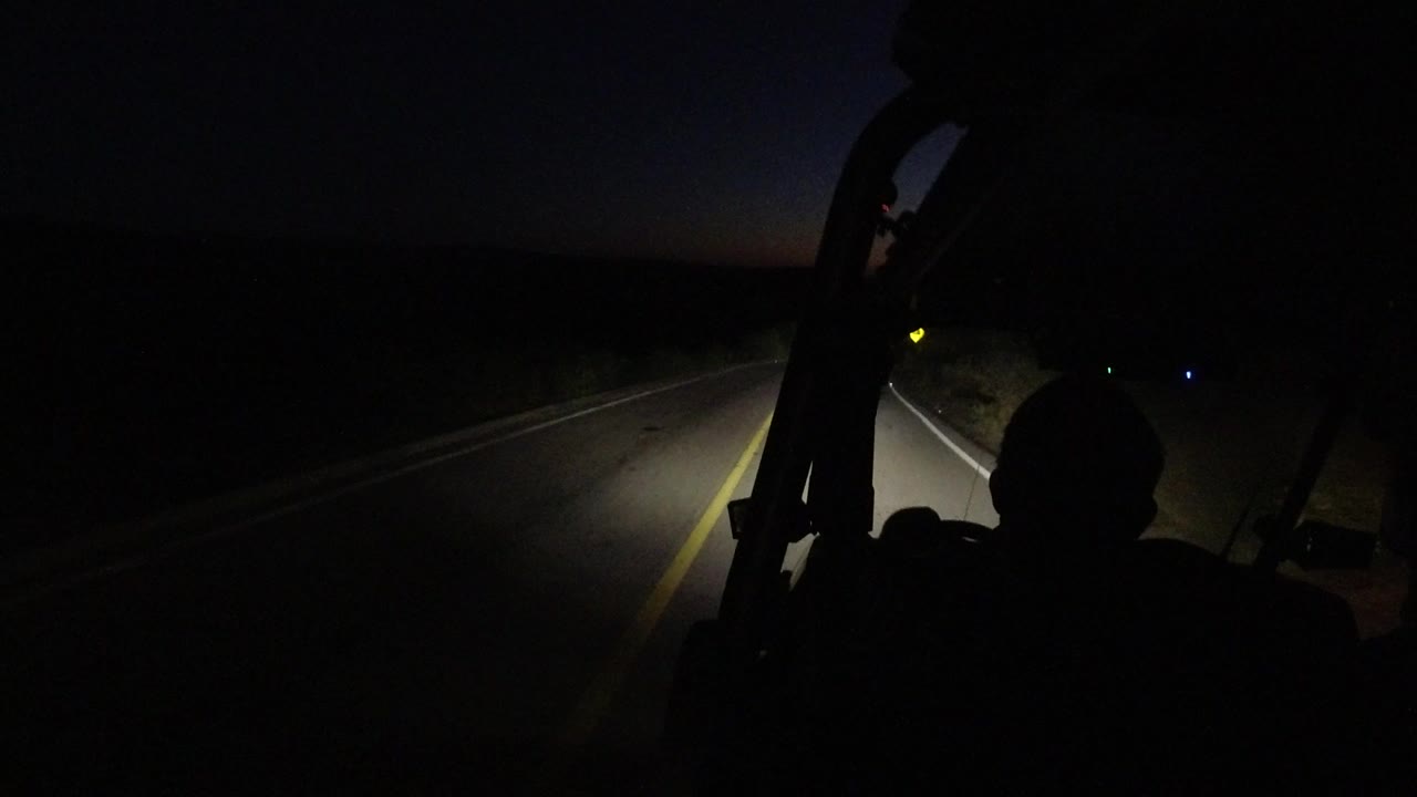 Dark in-car view in Los Cabos, Mexico, with a silhouetted occupant, windshield frame, yellow road line, and faint dawn color on the horizon.