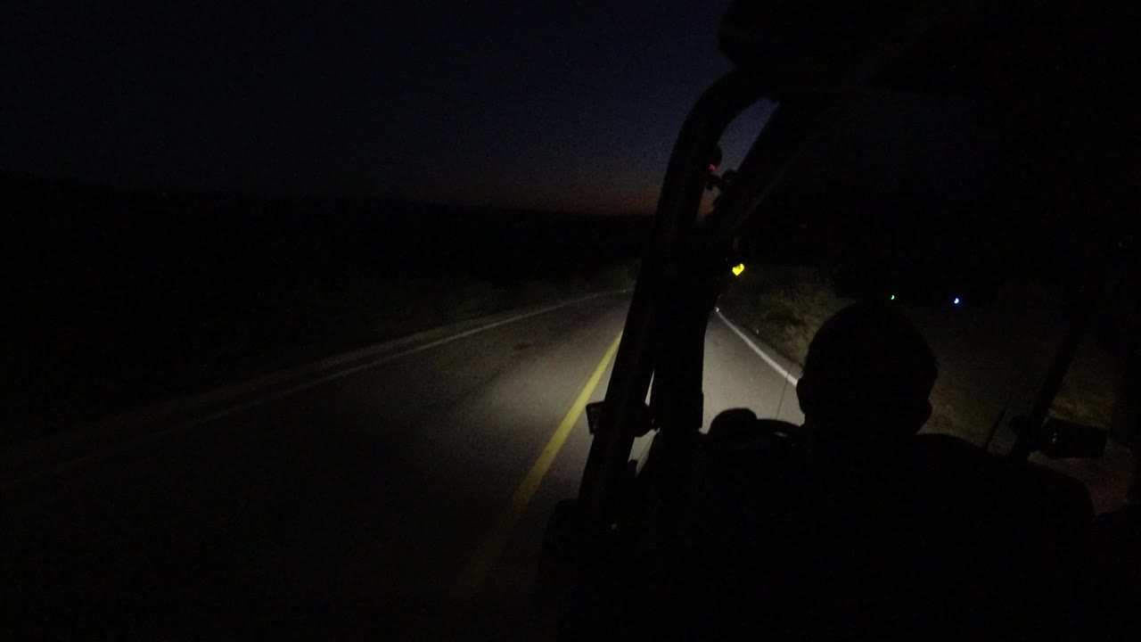 Inside-car view in Los Cabos, Mexico, with the road bending ahead, a dark passenger silhouette low in frame, and headlight spill across the lane edge.
