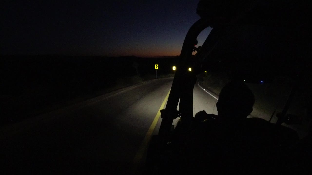 Opening frame of a Los Cabos road after sunset, with headlights lighting a curve, yellow chevron signs ahead, and a dark vehicle frame in the foreground.