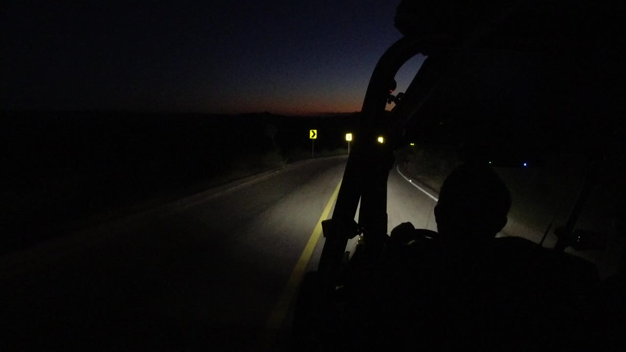 Los Cabos highway view at dusk with the headlight beam falling across the right lane, small roadside signs ahead, and a dark foreground silhouette inside the vehicle.
