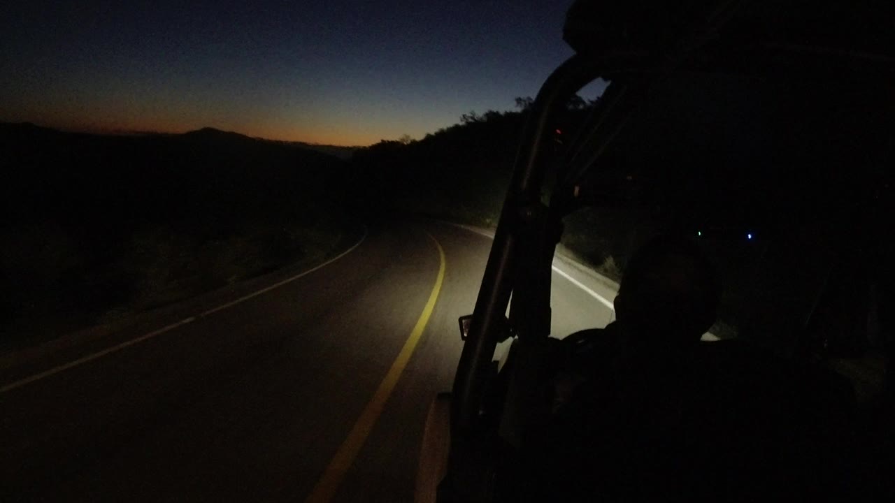 The Los Cabos road bends through darkness, with the yellow line catching the headlights and a dark occupant silhouette beside the vehicle frame.