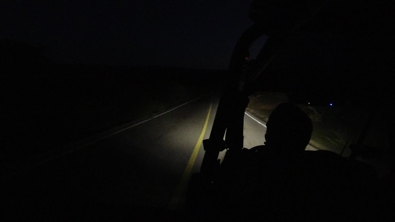 At 0:01, the mounted view remains very dark as the headlight beam catches the road edge and yellow line beside the silhouetted passenger in Los Cabos, Mexico.