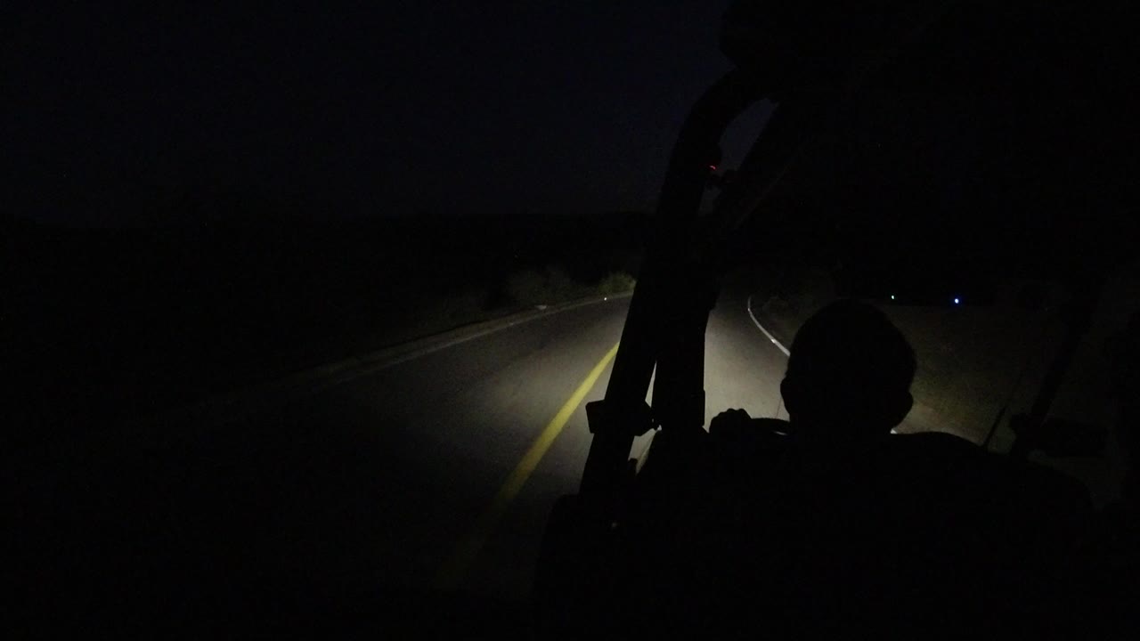 At 0:00 in Los Cabos, Mexico, a silhouetted driver sits in the foreground while headlights reveal a narrow stretch of dark asphalt and a yellow road line.
