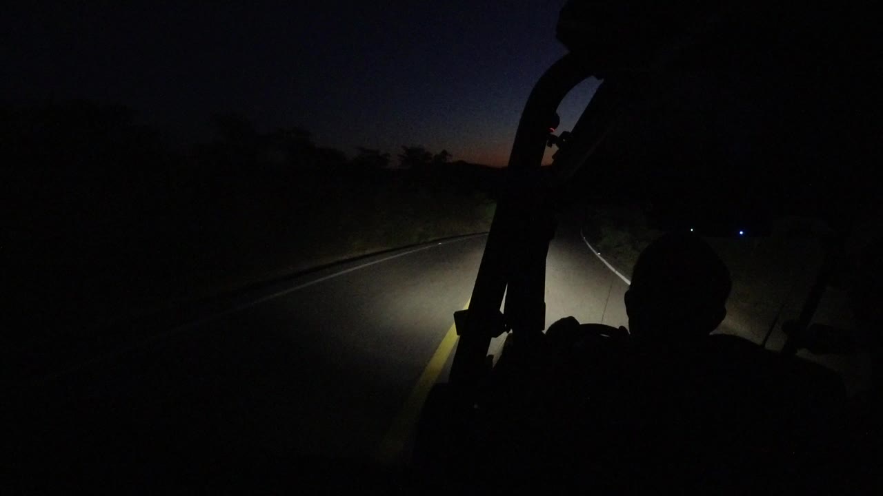 Forward view from inside a Jeep in Los Cabos, Mexico, with a roll-bar silhouette, headlights on the road, a faint horizon, and no visible people.
