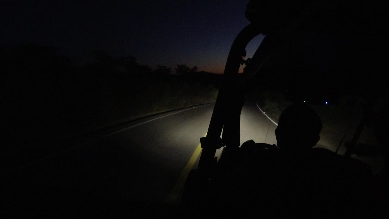 Interior Jeep view in Los Cabos, Mexico, with the road curving through darkness, brush at the roadside, small distant lights, and no visible people.