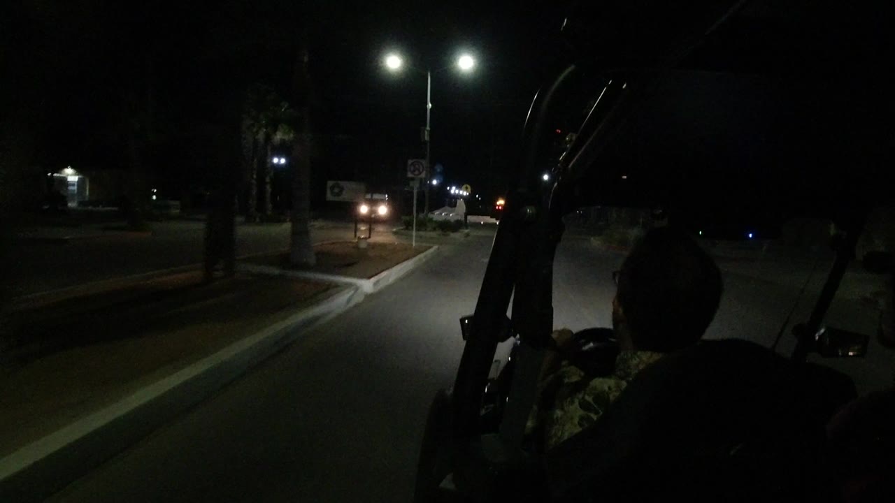 Los Cabos night street view from inside or behind an open vehicle, with a dark passenger silhouette at right and bright streetlights over the road ahead.