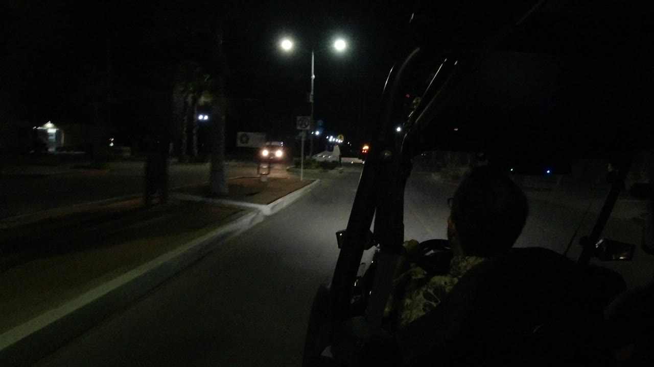 A nighttime Los Cabos roadway is framed by the vehicle windshield supports, with a seated passenger in silhouette and oncoming headlights near the streetlights.