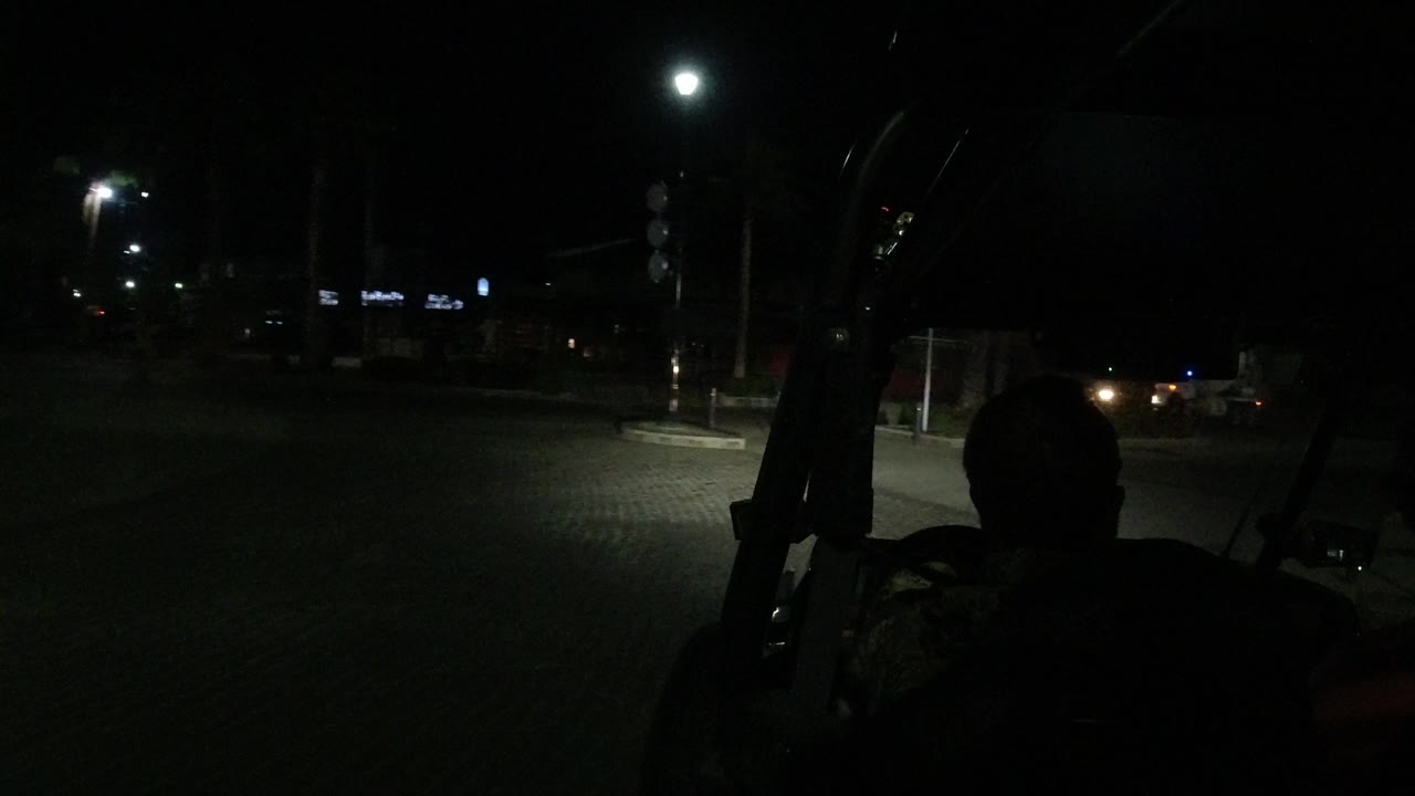 At 0:01 in Los Cabos, Mexico, the driver remains in silhouette as the vehicle faces a dim intersection with streetlights, a curb island, traffic signs, and distant building signs.