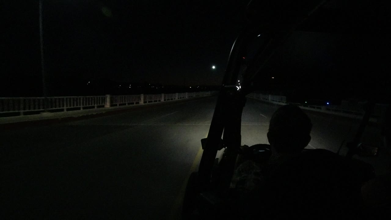 The Los Cabos road continues under a dark sky, with the rider's head and shoulders in silhouette, a roadside railing, and a small bright light ahead.