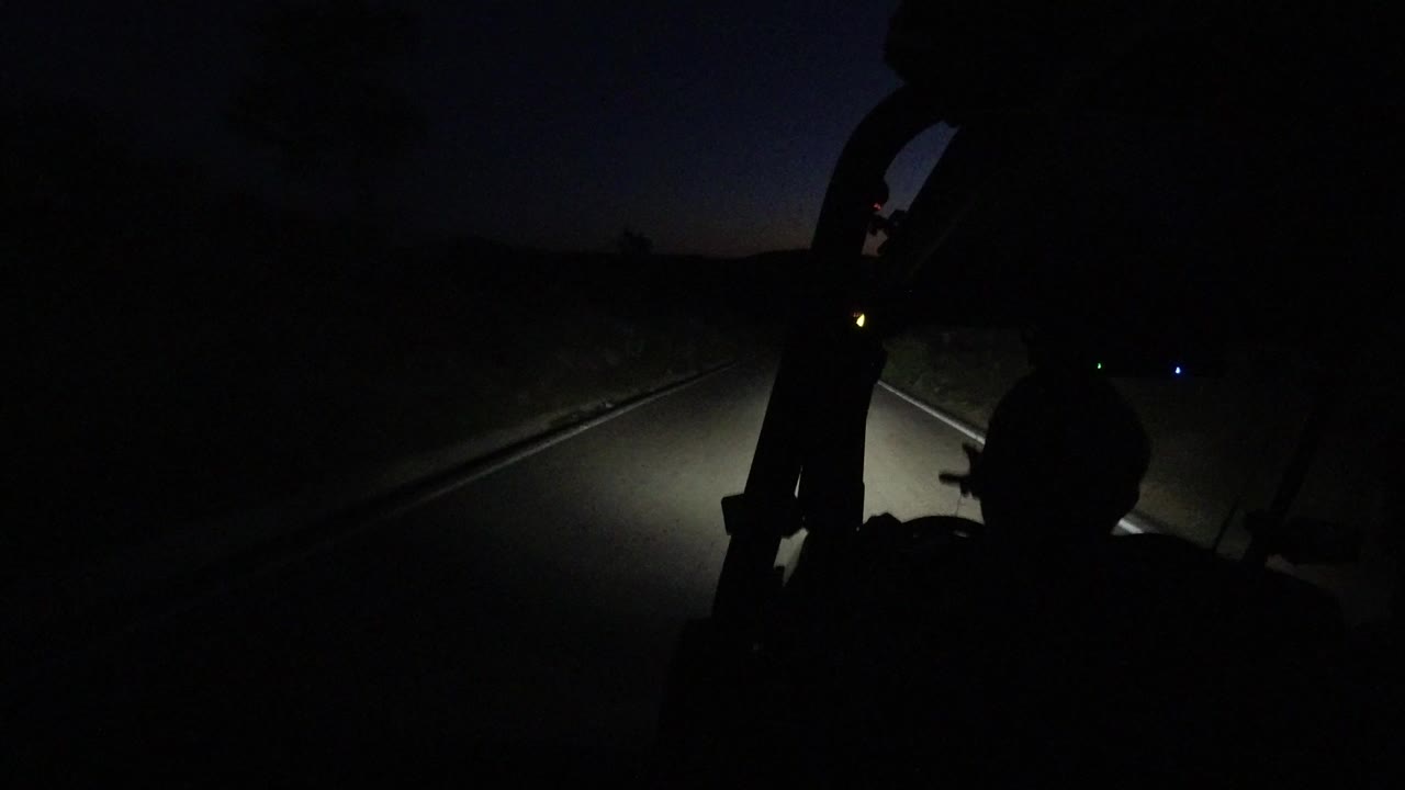 Opening frame of a dark paved road in Los Cabos, Mexico, with vehicle headlights illuminating the lane and a faint orange-blue horizon beyond the roadside.