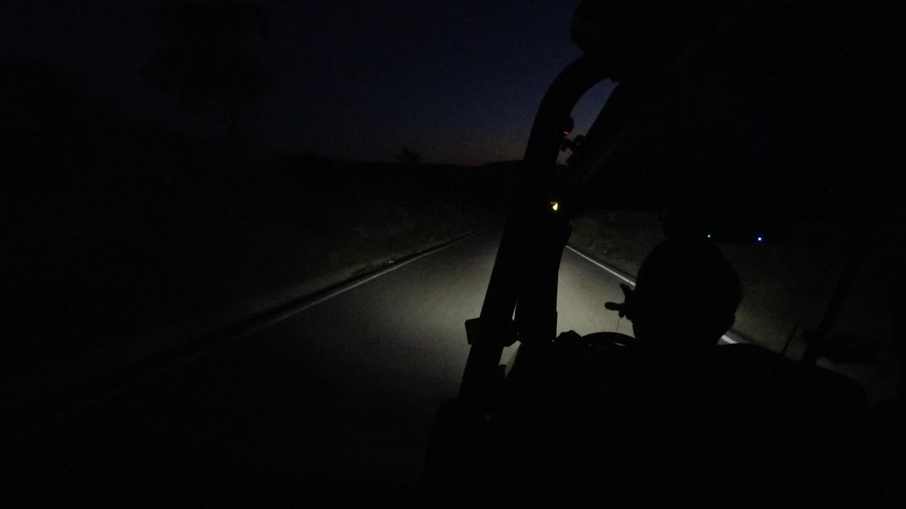 A forward road view in Los Cabos, Mexico, shows headlight spill on the pavement, darkness around the road edges, and a small warm point of light near the horizon.