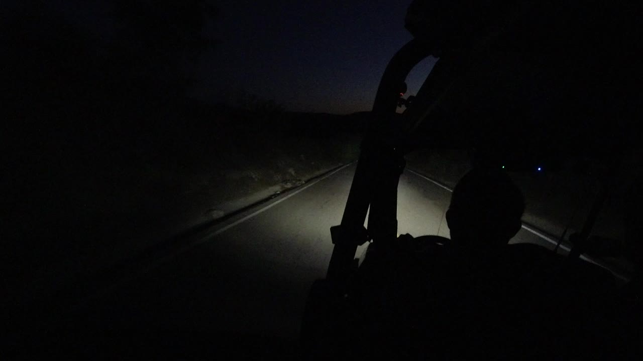 Night view from inside a vehicle in Los Cabos, Mexico, with a passenger silhouette at lower right and headlights illuminating the road ahead.