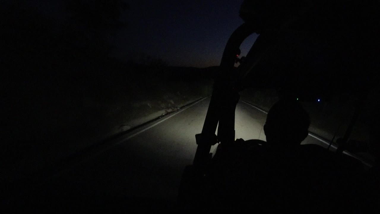 Inside-vehicle view in Los Cabos, Mexico, with the road curving through darkness and a seated passenger shown only as a black silhouette.